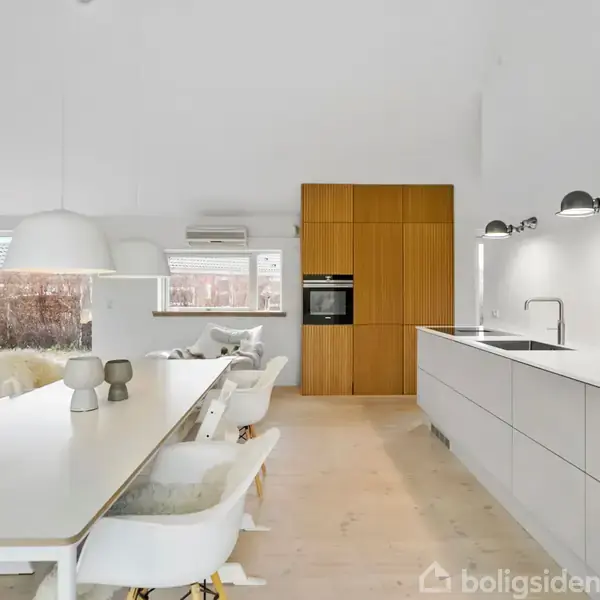 Et moderne køkken-alrum med hvidt spisebord og stole til venstre, lysekrone ovenover. Højre side har et hvidt køkkenbord med vask og lamper over. Baggrund har indbygget ovn og træskabe.