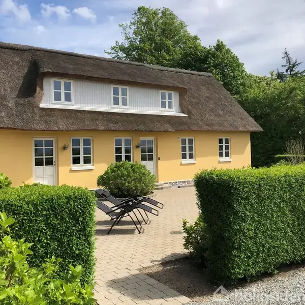 En gul, stråtækt hus bygning står i en haveomgivelser med frodige hække. Foran huset er der flisebelægning med to sorte liggestole og en lille busk.