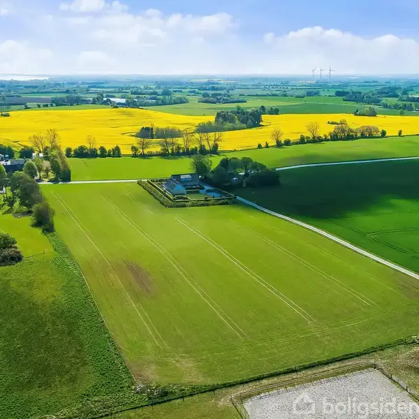 En gård med grønne marker og bygninger set oppefra. Gården er omgivet af marker og træer i et landligt landskab med en gul rapsmark i baggrunden.