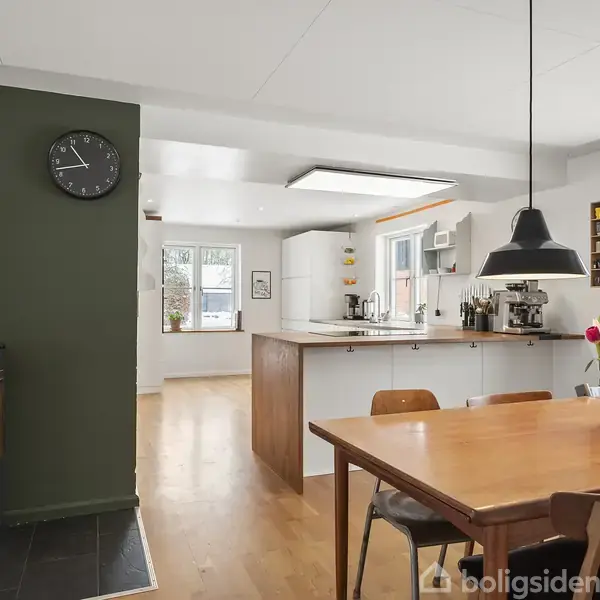 En spiseplads med et træbord og stole er i fokus, omgivet af en moderne åben køkkenindretning. En pejs brænder i hjørnet, og friske blomster dekorerer bordet.