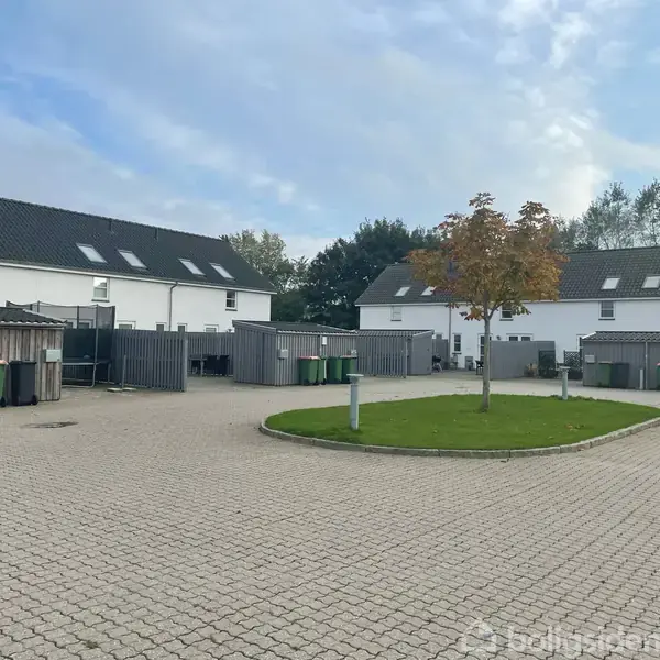 To hvide rækkehuse med sorte tage står over for hinanden. Foran er en belagt gårdsplads med grønne affaldscontainere og et lille centralt græsareal med et træ, omgivet af buske.