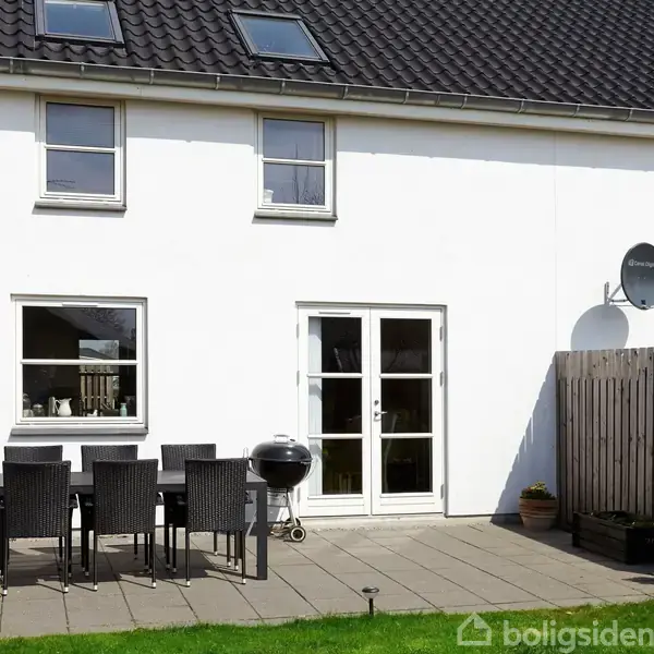 Hvidt rækkehus med sort tag, bord og stole på en terrasse, grill ved siden af. Træhegn og grøn græsplæne i forgrunden. Satelit skål med tekst "Canal Digital" på væggen.