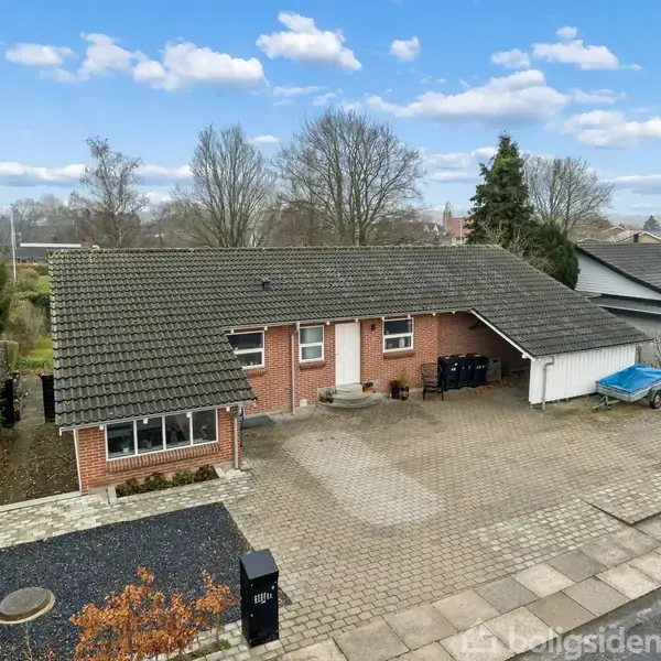 En murstensbungalow står stille på en flisebelagt indkørsel ved en villavej. Baggrunden prydes af høje, bladløse træer og omkringliggende haver. Postkassen er markeret med tallet "165".