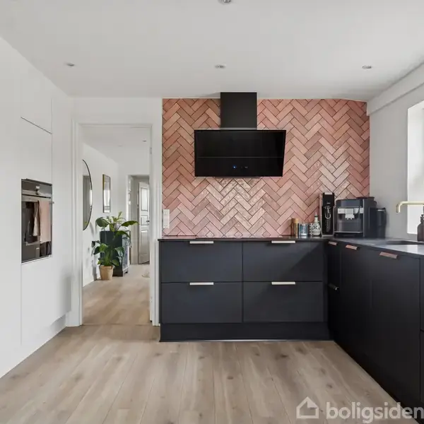 Køkken med sorte skabe står stille. Der er en orange murstensvæg som baggrund. Rummet har trægulv, et stort vindue og diverse køkkenredskaber på bordet.