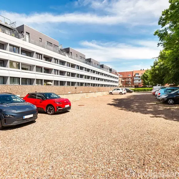 Biler holder parkeret på en grusplads foran en moderne, lang hvid og grå boligbygning. Solen skinner og et stort træ står til højre.