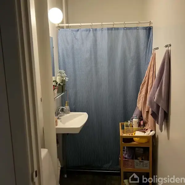 En håndvask hænger på væggen i et lille badeværelse. Foran hænger et blåstribet bruseforhæng. Til højre hænger håndklæder, og der er en gul opbevaringsvogn fyldt med toiletartikler.