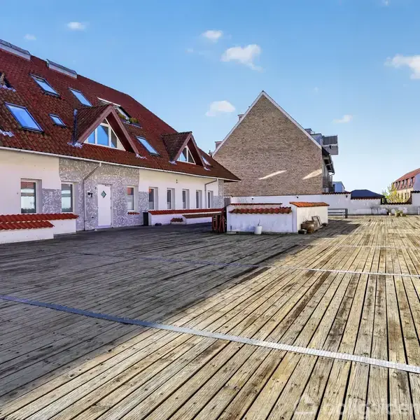 Bygningens tagterrasse med træplanker strækker sig bredt ud. Ved siden af er der rødt tegltag med tagvinduer, hvor solen skinner. Baggrund viser flere bygninger under en klar blå himmel.