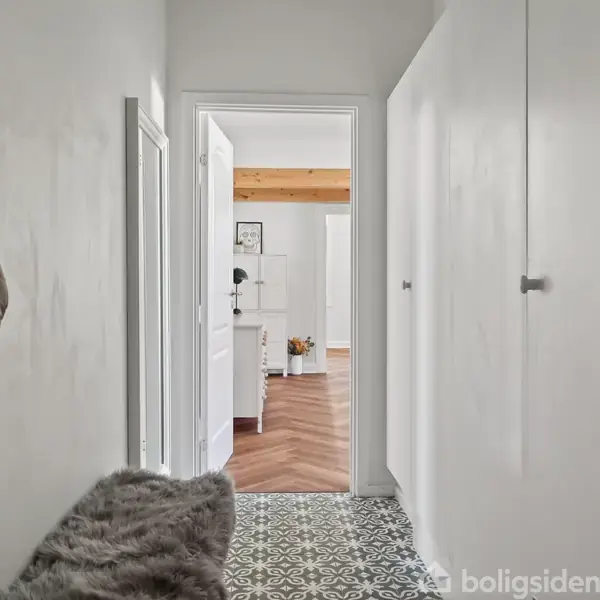 Gang med flisegulv, hvor en jakke hænger på væggen. I baggrunden ses et rum med trægulv og møbler. Væggene er hvide med skabe på højre side.