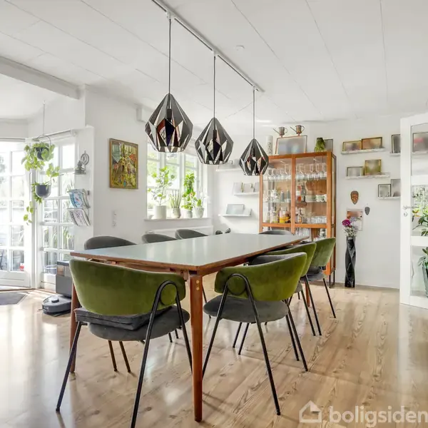 Spisebord med seks grønne stole omringet af moderne lamper i en lys stue med trægulv, glasdøre, planter, og et skab fyldt med glas.