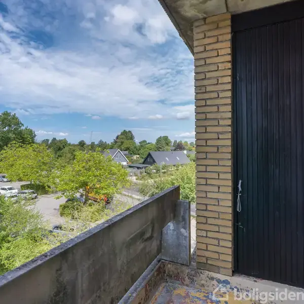 Altan med udsigt over et grønt boligområde. Kalkstensvægge og sort dør er synlige i højre side. Træer og et hus kan ses i baggrunden under en skyet blå himmel.