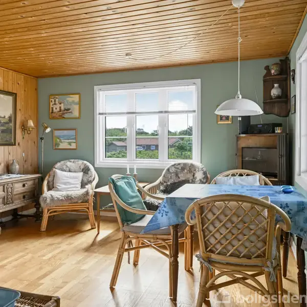 En hyggelig stue med et spisebord dækket af en blå dug. Rattan-stole står omkring bordet. Trægulv og -loft giver et varmt miljø. Vindue med udsigt til have omgivet af malerier.