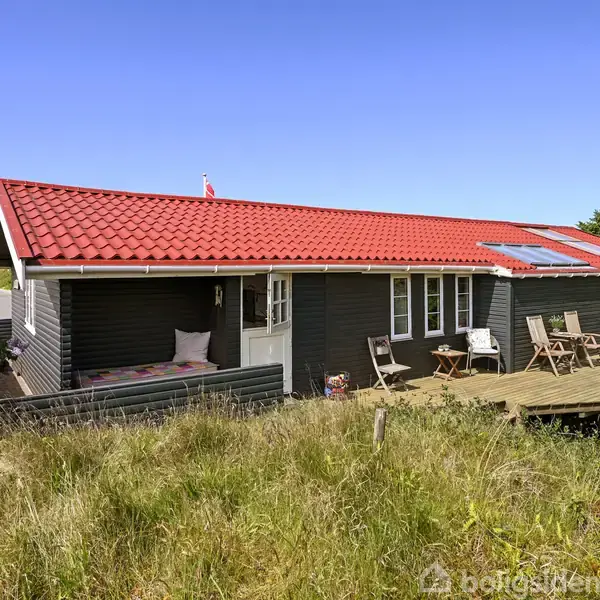 Sommerhus med rødt tegltag og mørkegrå facade ligger i en naturpræget have. En åben træterrasse har bord og stole, omgivet af grønne buske og vilde græsser under en blå himmel.
