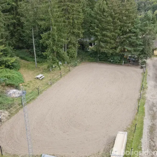 En sandridebane omkranset af et hegn omgivet af tætte træer. Til højre ses en bygning med grønne tage. Stier leder rundt i området.