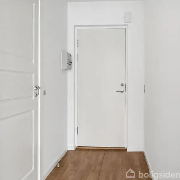 Hvid dør står lukket i en smal, hvid gang med trægulv. Gangens vægge er tomme, og der ses et enkelt gråt vægpanel. Ingen tekst er synlig.