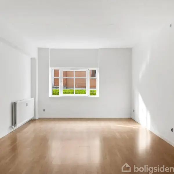 Et tomt værelse med trægulv. Et åbent vindue giver udsigt til en murstensbygning udenfor. En radiator er placeret til venstre. Boligsiden-logo nederst til højre.