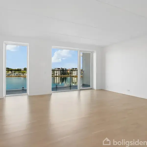 Et tomt rum har trægulv og hvide vægge. Gennem store vinduer ses en balkon med udsigt til en marina eller havneområde i sollys.