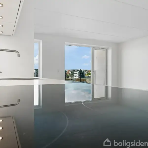 Moderne køkken med en blank sort bordplade reflekterer en vandhane; store vinduer i baggrunden giver udsigt til en by og lys strømmer ind i det lyse rum.