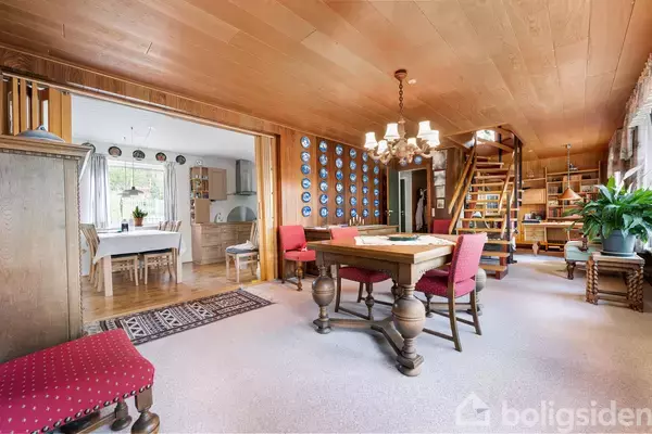 Et stueområde med trægulv og loft, har et spisebord omgivet af røde stole. Til højre en trætrappe og reol. I baggrunden ses et køkkenområde med spisebord.