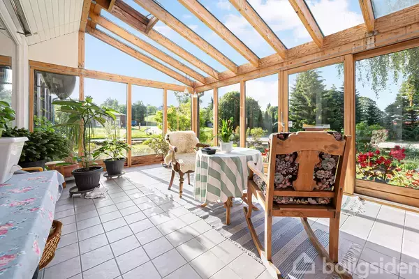 Et lyst vinterhaveområde med tæppebelagte stole om et bord med en ternet dug. Store vinduer og åbent til en grøn have med blomster og træer i baggrunden.