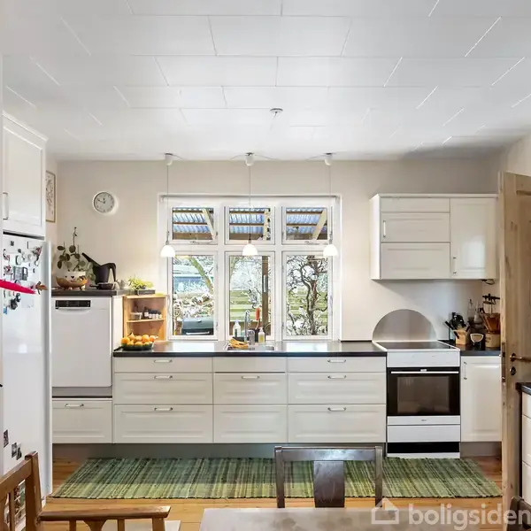 Et lyst køkken med hvide skabe og skuffer, bordplader med køkkenartikler, og et spisebord i forgrunden. Et vindue viser en have i baggrunden. Ingen tekst i billedet.