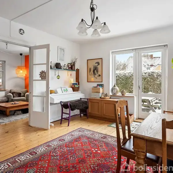 Et lyst rum med træmøbler og orientalsk tæppe har åbne franske døre til en stue med sofaer. Vinduer viser sneklædt have udenfor. Rummet er dekoreret med planter og billeder.