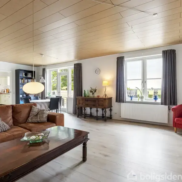 Stue med brun lædersofa og sofabord. Sofaen har puder. Store vinduer med gardiner bagved giver lysindfald. Rød stol i højre hjørne; spisestue og køkken i baggrunden.