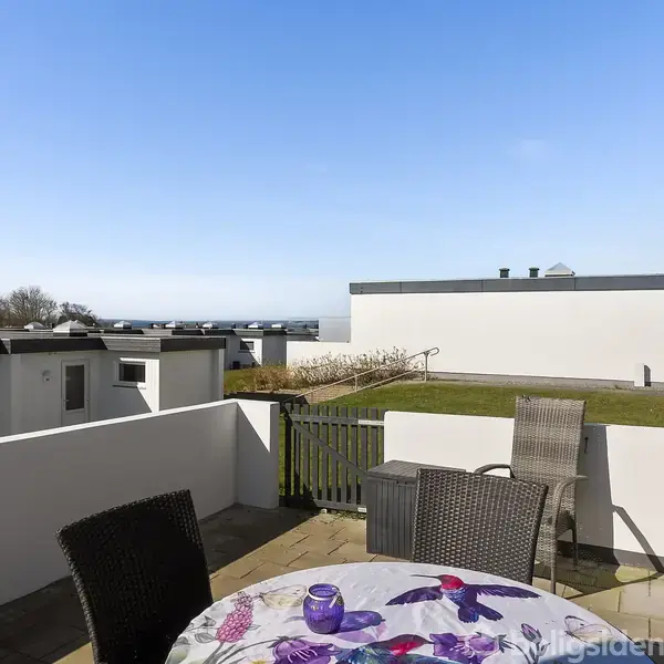 Rækkehuse ligger stille i en grøn mark tæt ved kysten med udsigt over havet. I baggrunden ses en lille havn og flere huse langs kysten.