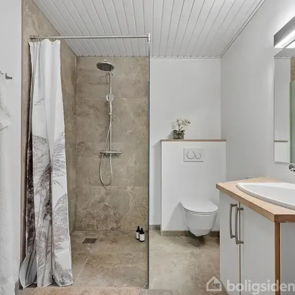 Badeværelse med bruser. Brusekabine med glasvæg, beige fliser og brusehoved til venstre. Til højre håndvask med spejl, toilet, og dekorativ plante. Håndklæder hænger på væggen.