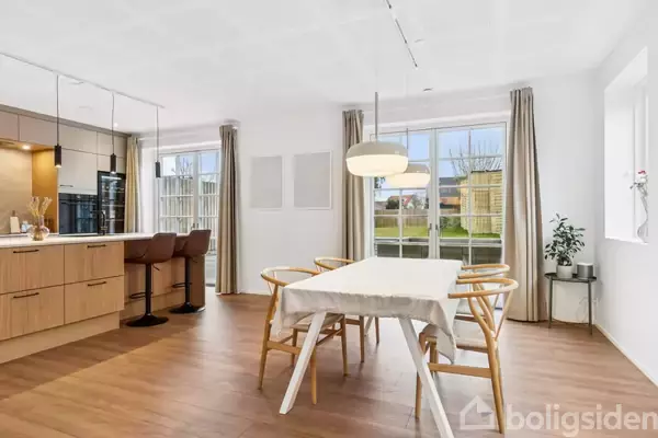 Spisebord dækket med dug står i lys spisestue. Stole omkranser bordet. Store vinduer med udsigt til have. Køkken med barstole og skabe er synligt til venstre.