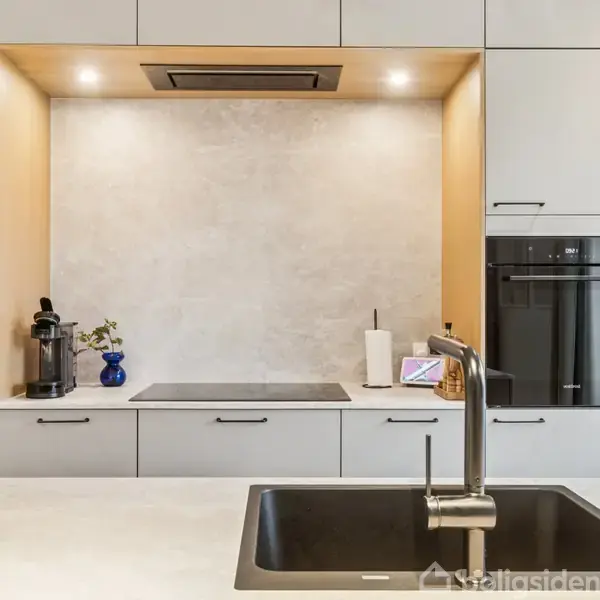 Køkken har en vask i forgrunden, kogeplade og kaffemaskine i midten, og moderne hvide skabe samt indbygget ovn i baggrunden.