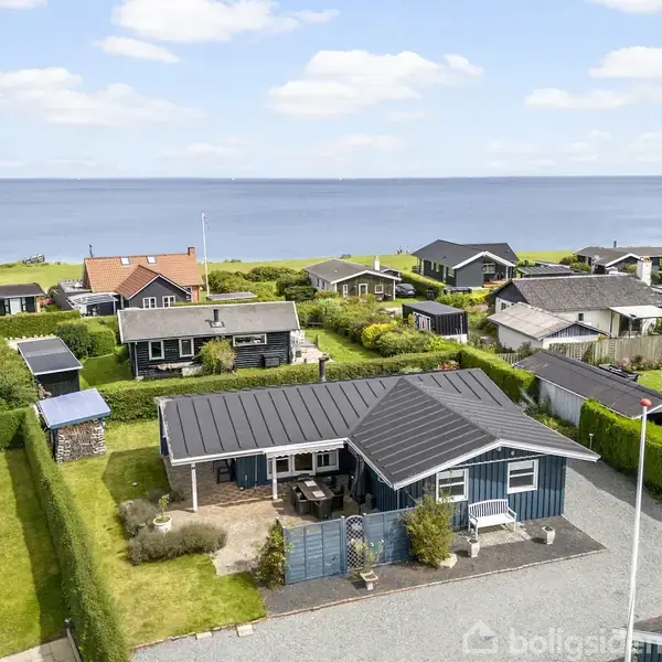 Et lille hus med sort tag og blå facade står stille i et parcelhusområde nær vandet. Omgivet af velplejede haver, med en klar udsigt til havet i baggrunden.