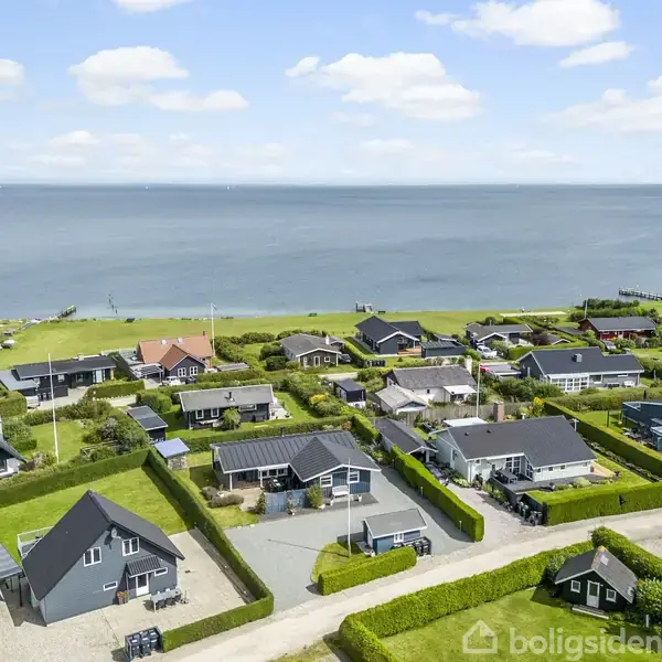 Huse ligger tæt samlet i et kystnært område med havudsigt under en blå himmel med spredte skyer. Græsarealer omgiver ejendommene, og havet strækker sig i baggrunden.