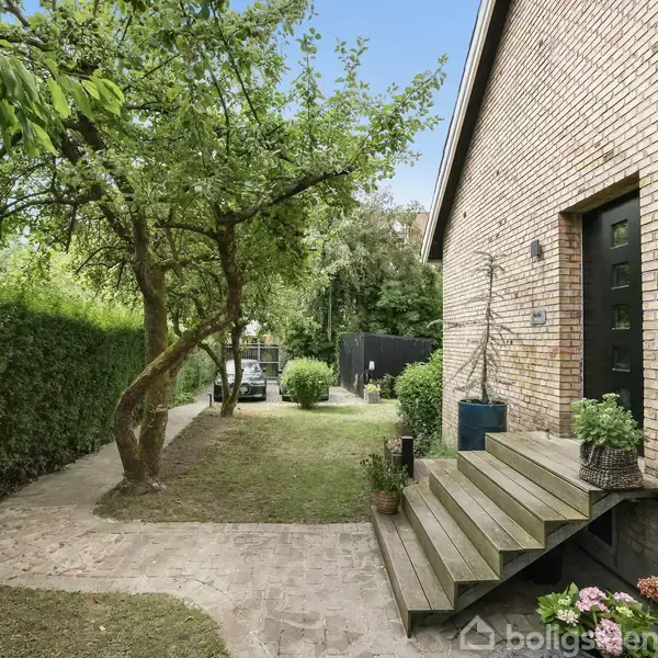 Et murstenshus med en sort dør og trappeopgang. Foran huset er der planter i krukker. En snoet flisegang og en lille have med et træ omgiver huset.