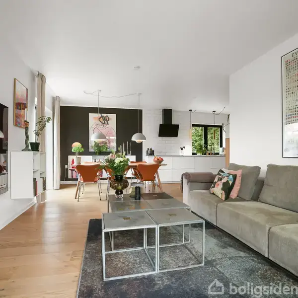 Stue med grå sofa, sofabord og tæppe i centrum. I baggrunden ses spisebord med orange stole, planter og kunst på væggene. Rummet har hvidmalede vægge og trægulv.