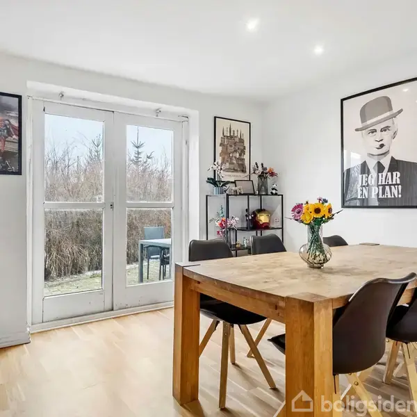 Et spisebord med blomster i en vase står i et lyst rum med trægulv. Der er fire stole omkring bordet. På væggen hænger en plakat med teksten "Jeg har en plan". I baggrunden ses en glasdør, der fører til en terrasse omgivet af buske.