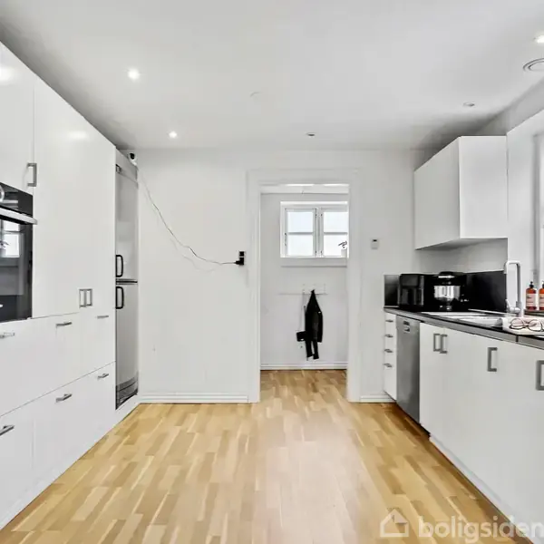 Et moderne køkken med hvide skabe og trælaminatgulv. Et komfur og ovn er indbygget til venstre. Store vinduer til højre giver naturligt lys. En sort jakke hænger på en krog.