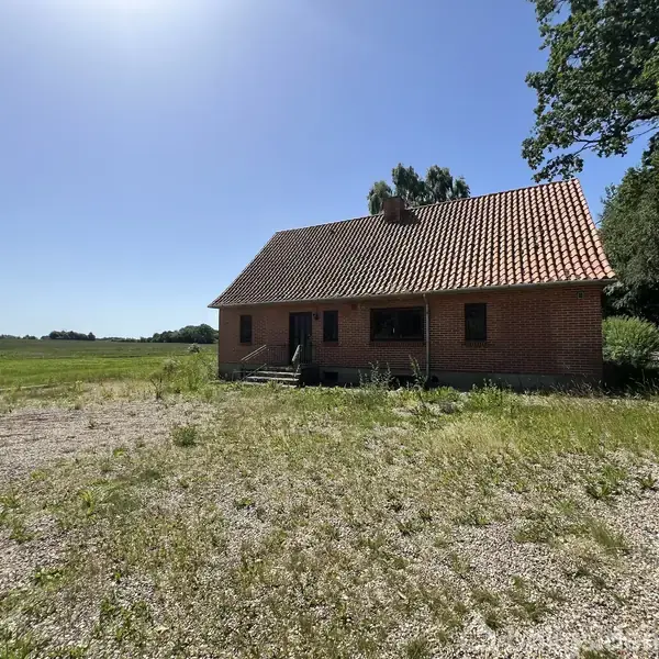 Et rødt murstenshus med rødt tegltag står midt i en åben, solrig græsfyldt mark, omgivet af træer og en blå himmel.