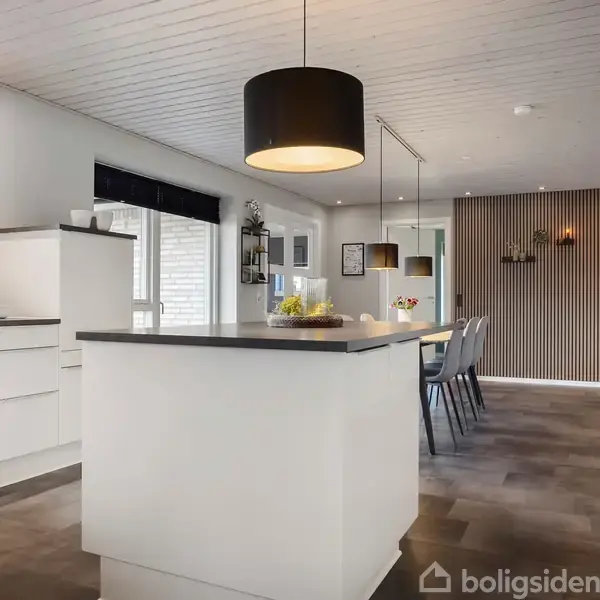Køkken med hvidt interiør, stor central køkkenø, og sort lampe. Spisebord med stole i baggrunden. Væggene er lyse med et stribet panel. Gulvet er gråt.