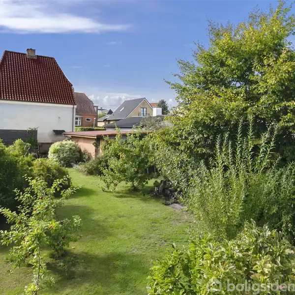 Et hus med rødt tag står roligt i en frodig have med grønne buske og træer. I baggrunden kan man se flere huse og en klar blå himmel.