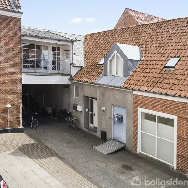 En gårdhave med cykler parkeret foran en murstensbygning. Bygningen har rødt tegltag og et lille gavlvindue. Der er også en skydedør og flere postkasser.