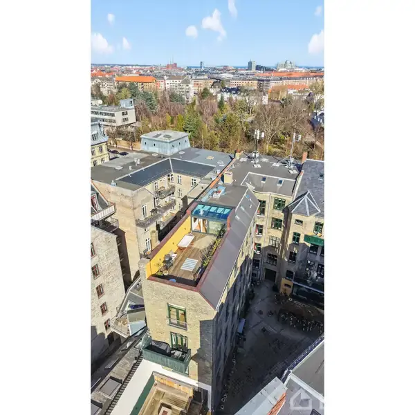 Bygning med tagterrasse har møbler og planter. Terrasseaktivitet antyder socialt samvær. Omgivet af bylandskab med flere bygninger og grønne områder i baggrunden under blå himmel med skyer.