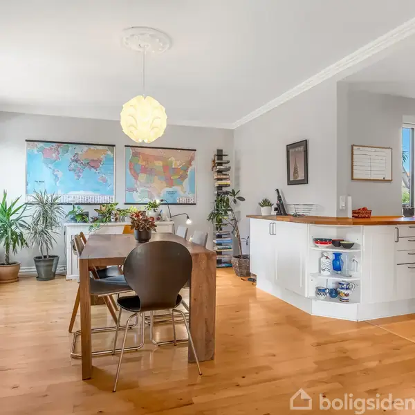 Spisebord med stole står i lys spisestue med træplankegulv. Planter og verdenskort pryder væggene. Åben køkkenø til højre, omgivet af lysegrå vægge og store vinduer.