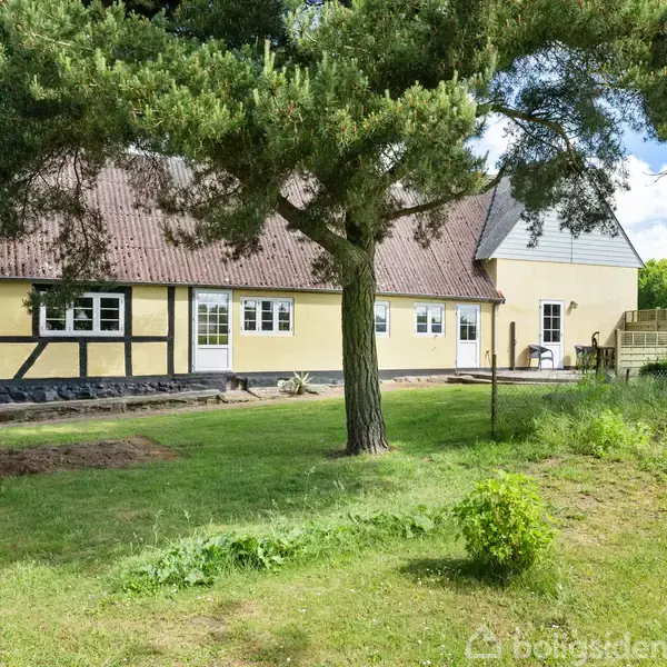 Et gult bindingsværkshus med rødt tag står i en grøn have med træer og græs omkring. Huset har hvide vinduer og en lille terrasse. Boligsiden-logo nederst i billedet.