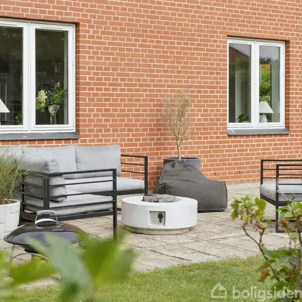 Terrasse med grå sofasæt og hvid ildsted, omgivet af grønne planter i potter. Baggrunden er en murstensvæg med to vinduer. Fliser dækker jorden, og buske omkranser området.