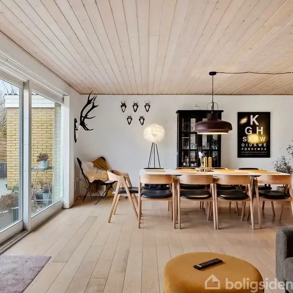 Spisestue med træbord og seks stole, dekoreret med vægkunst og en gulvlampe. Stort vindue med udsigt til terrasse med planter. Vandrette træpaneler i loftet.