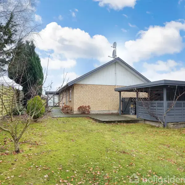 En gulstensbygning med et gråt tag står i en frodig have. Ved siden af er en overdækket terrasse af træ. Træer og buske omgiver græsplænen, under en delvist skyet himmel.