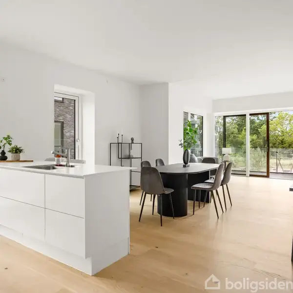 Et moderne køkken-alrum med en hvid køkkenø, spisestuebord og stole. Store vinduer til venstre giver udsigt til en grøn have. Indretningen er minimalistisk og lys.