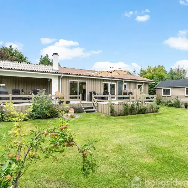 Et enkelt hus med terrasse står i en velplejet have med grøn græsplæne og frugttræer. Ved siden af huset ses et lille skur, og himlen er klar med få skyer.