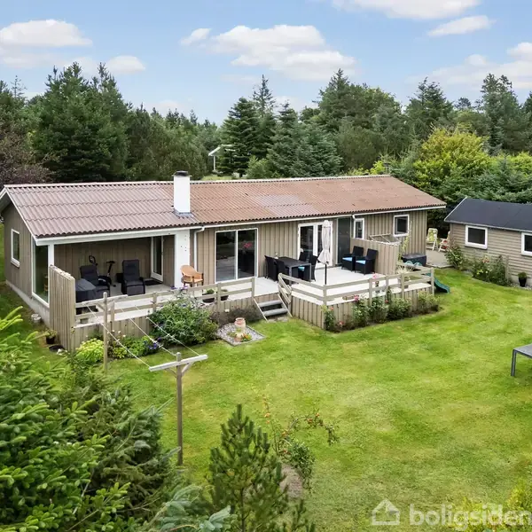 Et enfamiliehus med en træterrasse står i en frodig have omkranset af træer. Ved siden af huset er et mindre skur. Græsplænen er velplejet med udendørsmøbler.