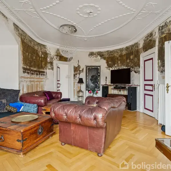 En stue med møbler i en afslappet opsætning. To læderlænestole og en sofa omkranser et træbord. Væggene viser afskallet maling, og der hænger en lampe fra loftet. En fladskærms-TV er monteret på væggen.